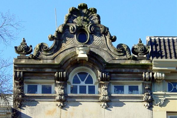 Verhoogde lijstgevel in Lodewijk XIV-stijl met attiekvormige versiering met kuif, bovenop de halfcirkelvormige verhoging, en hoekvazen (© Walther Schoonenberg)